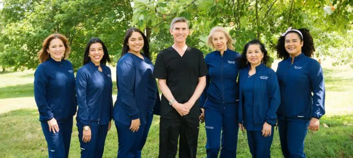 Dental team in blue scrubs posing outdoors under trees.